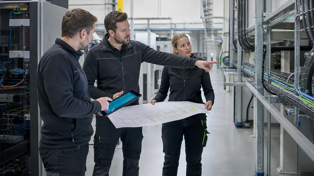 Three specialists compare an infrastructure plan with a tablet and physical routes in a production environment